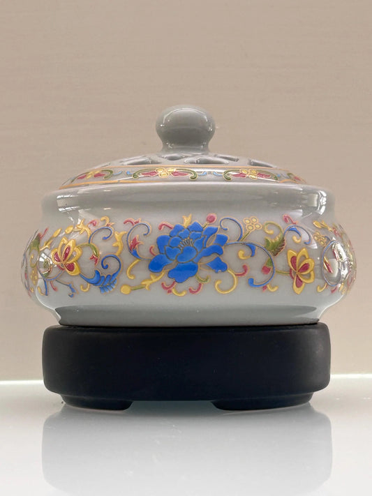 A white ceramic lidded box with colorful hand-painted floral patterns, resting on a black circular base against a neutral beige background.